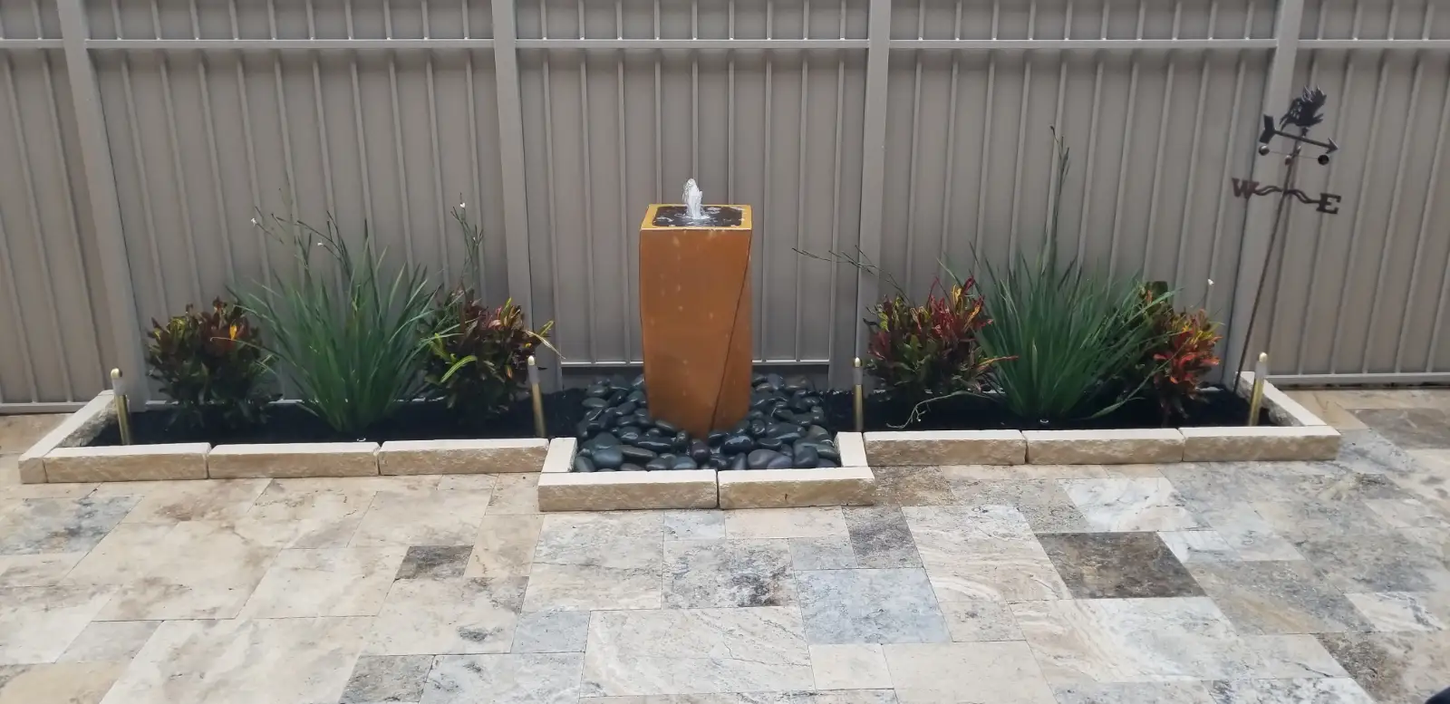 Travertine patio with a corten water fountain, river rock and croton beds